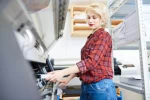 Junge blonde Frau beim Plotten in Berlin, die an einem großen Plotter in einem hellen Raum arbeitet. Sie trägt eine rote karierte Bluse und eine blaue Jeans.